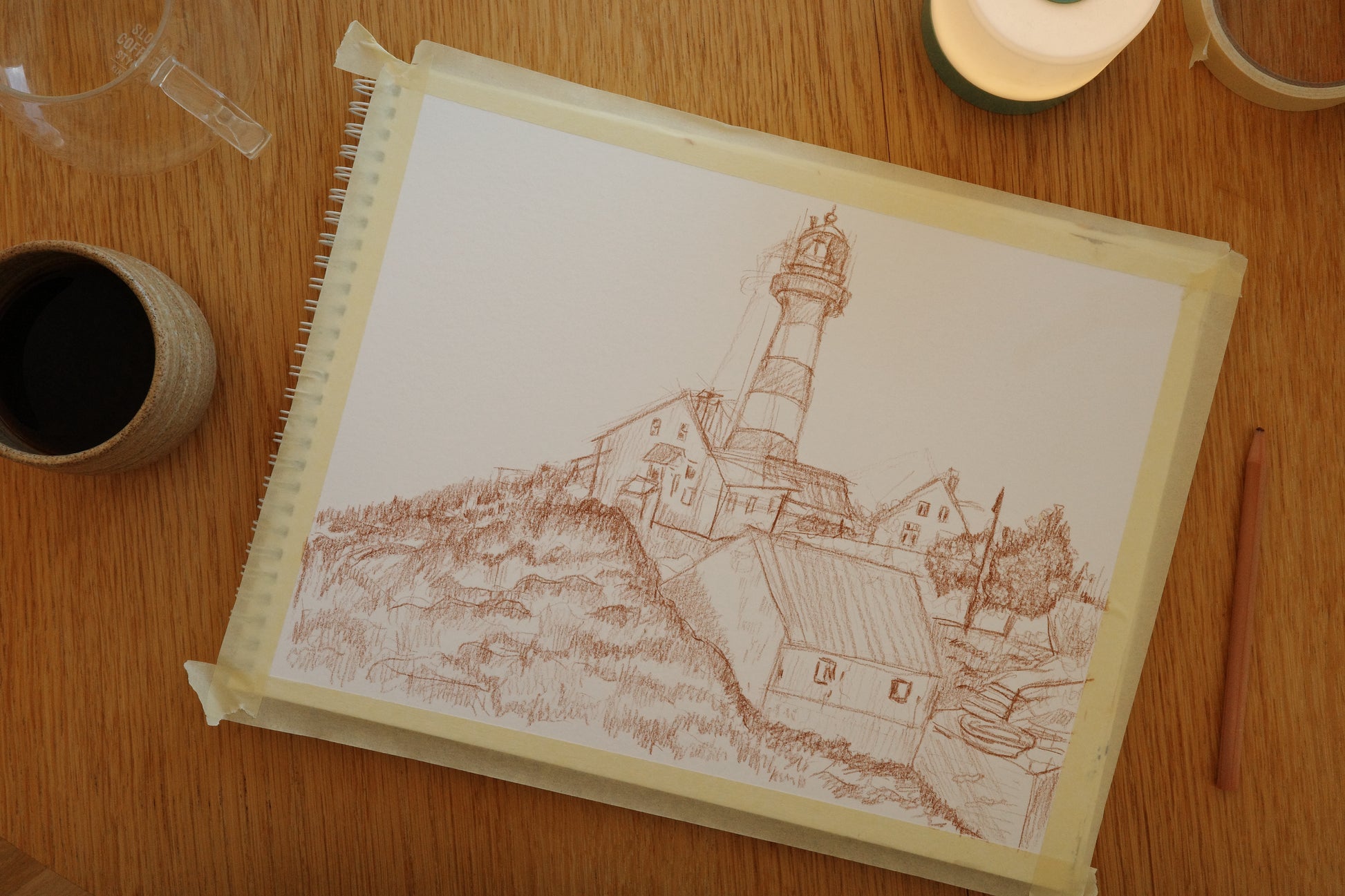 Spiral-bound notebook with a lighthouse drawing on a wooden surface with a cup and candle.